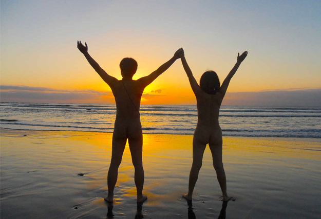 Village Naturiste du Cap d’Agde - Un homme et une femme nus sur une place de dos au coucher de soleil les bras écartées.