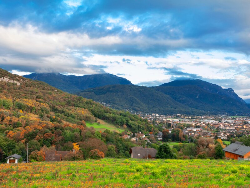 Massage érotique Chambéry - Vue panoramique sur Chambéry entourée de montagnes, avec des collines verdoyantes et des maisons sous un ciel nuageux.