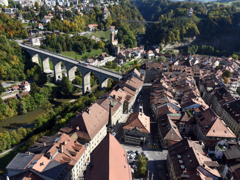 Massage érotique Fribourg - Vue aérienne de la ville de Fribourg avec un pont en arc traversant une vallée verdoyante, entourée de bâtiments anciens et de collines.