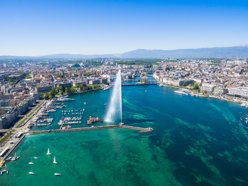 massage érotique Genève - Vue aérienne de Genève avec le Jet d'Eau et le lac Léman, entourés par la ville et les montagnes sous un ciel bleu clair.