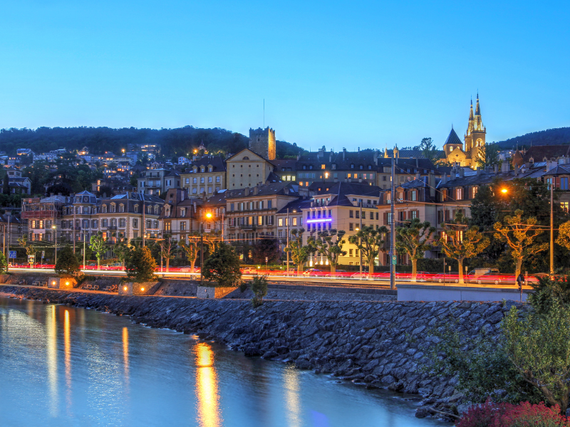 massage érotique Neuchâtel - Vue nocturne de la ville de Neuchâtel avec des bâtiments éclairés au bord du lac, sous un ciel clair.