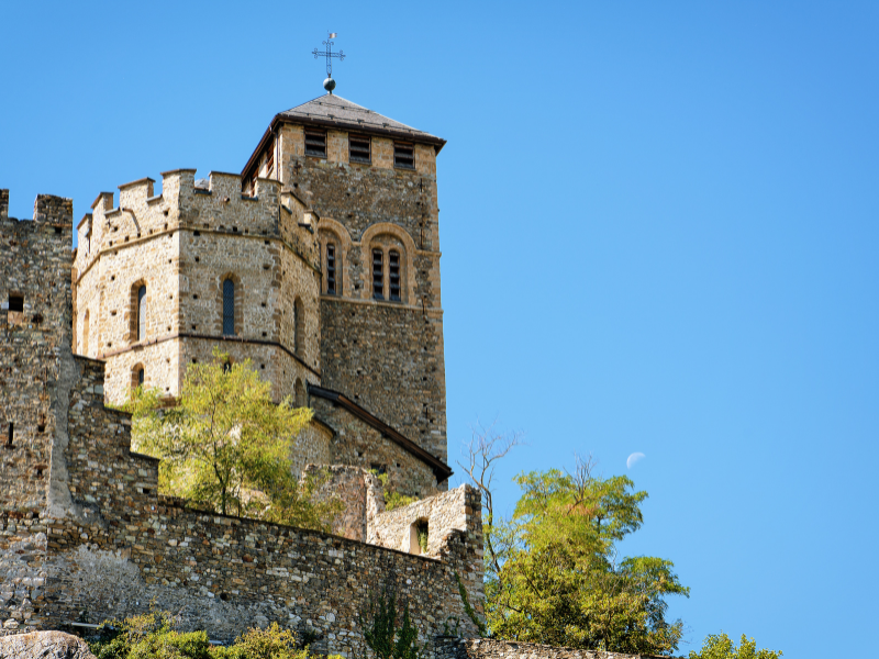 Massage érotique Sion - Vue d'une tour en pierre d'un château à Sion, entourée de végétation sous un ciel bleu clair