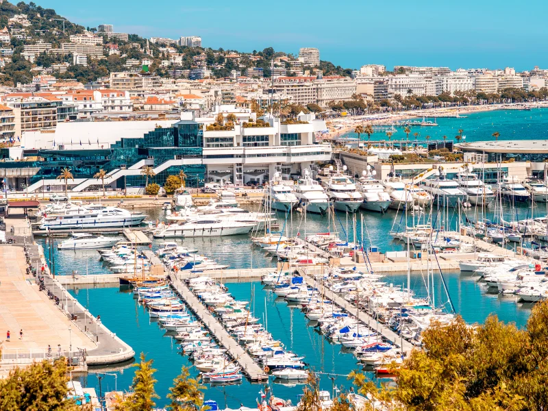 Massage érotique Cannes - Vue du port de Cannes avec ses yachts et bateaux amarrés, entouré de bâtiments modernes et de collines verdoyantes en arrière-plan.