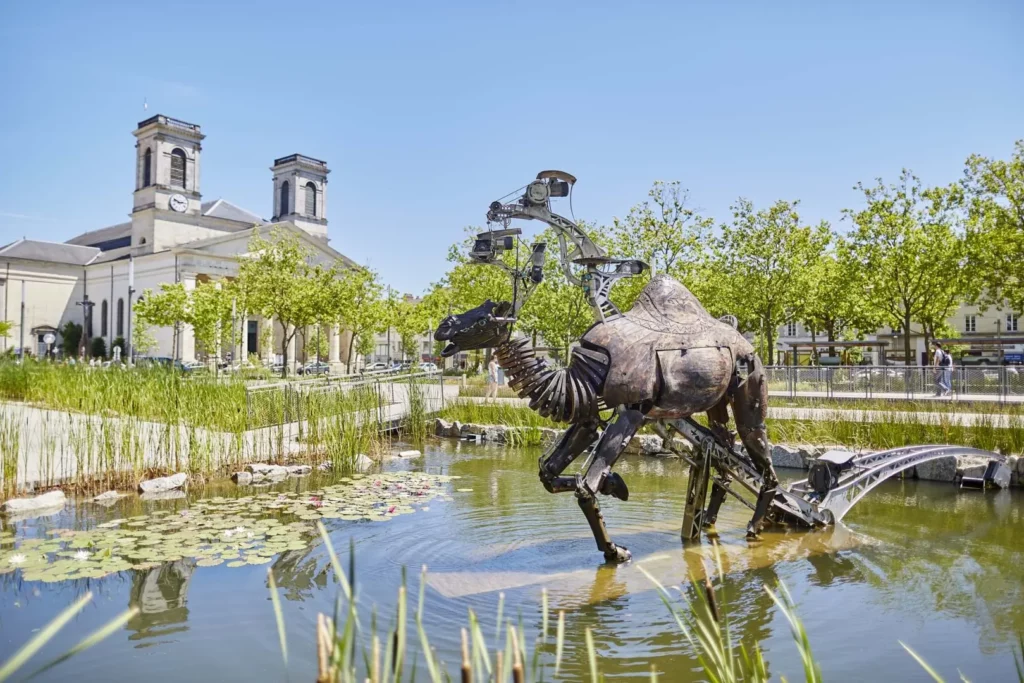 Massage érotique La Roche-sur-Yon : Sculpture mécanique d'un dromadaire dans l'étang de la place Napoléon à La Roche-sur-Yon, avec l'église Saint-Louis en arrière-plan sous un ciel dégagé.