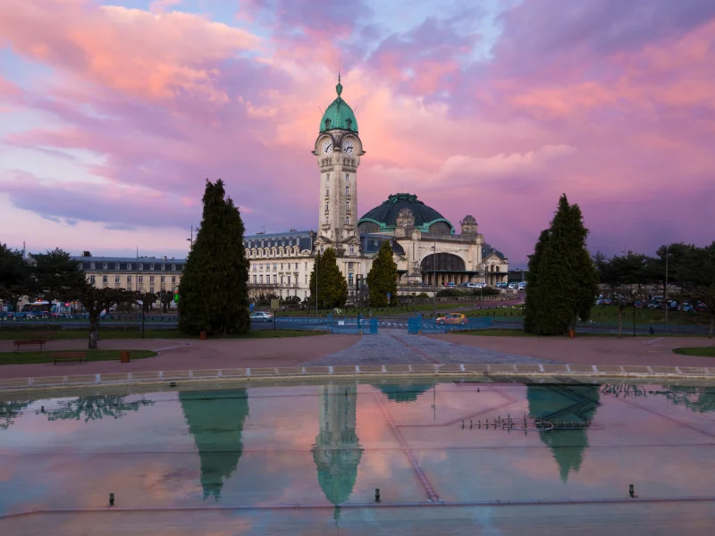 massage érotique Limoges - Vue du coucher de soleil sur la gare des Bénédictins à Limoges, avec ses reflets dans une fontaine au premier plan et un ciel coloré en arrière-plan.