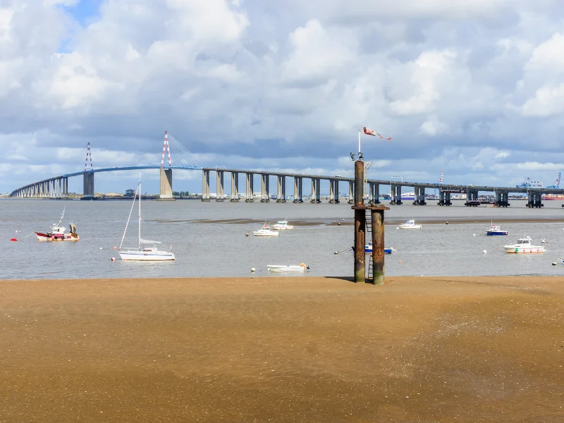 Massage érotique Saint-Nazaire - Vue sur le pont de Saint-Nazaire avec des bateaux amarrés dans l'estuaire de la Loire sous un ciel nuageux.